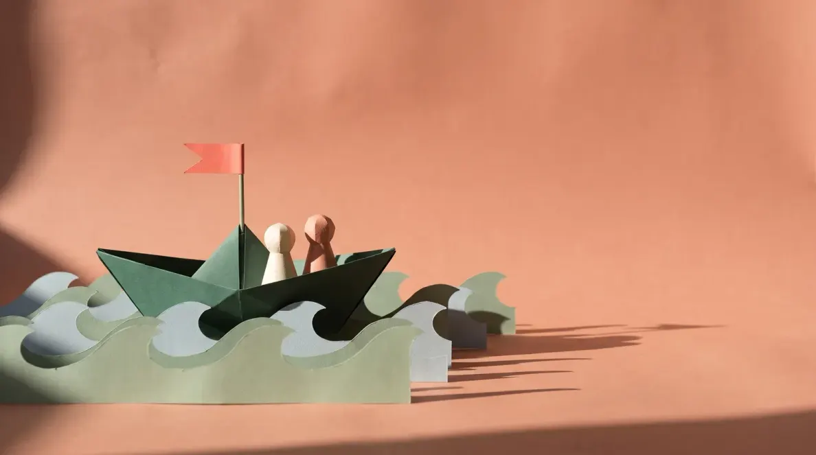 Ein gefaltetes Papierboot mit roter Fahne fährt über Wellen aus Papier, darin sitzen zwei Spielfiguren nebeneinander
